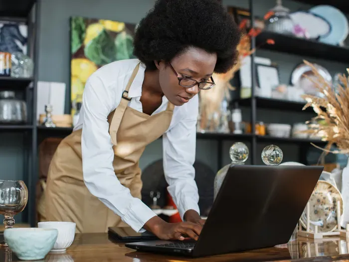 Shop owner in apron working on a laptop in a boutique, showing how CityBiz Network Hub helps manage local listings